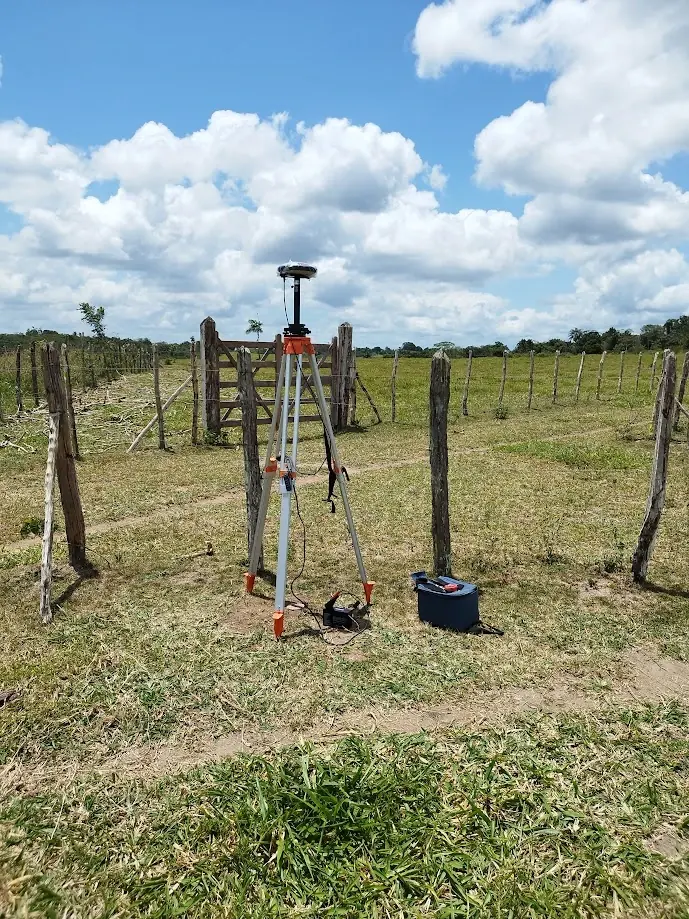 Receptor GNSS base em tripé laranja instalado numa pastagem cercada, com bateria externa ao lado