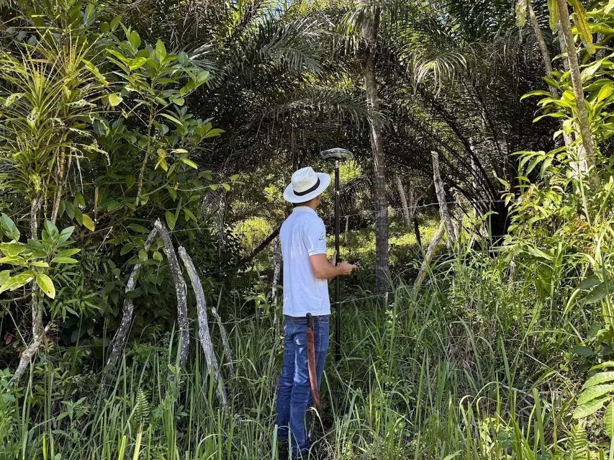 Ralph Araujo com receptor GNSS em cerca de divisa, cercado por vegetação densa e palmeiras