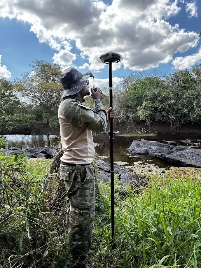 Técnico com chapéu e roupa camuflada operando receptor GNSS na margem de um rio com pedras escuras