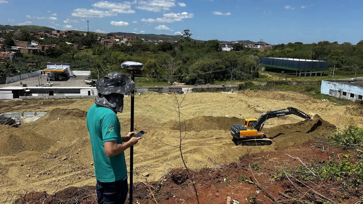 Técnico com receptor GNSS e coletora coletando pontos sobre um talude, com escavadeira trabalhando na área de terraplenagem ao fundo
