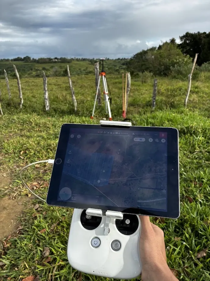 Controle remoto de drone com tablet exibindo plano de voo em faixas; ao fundo, receptor GNSS em tripé numa pastagem cercada